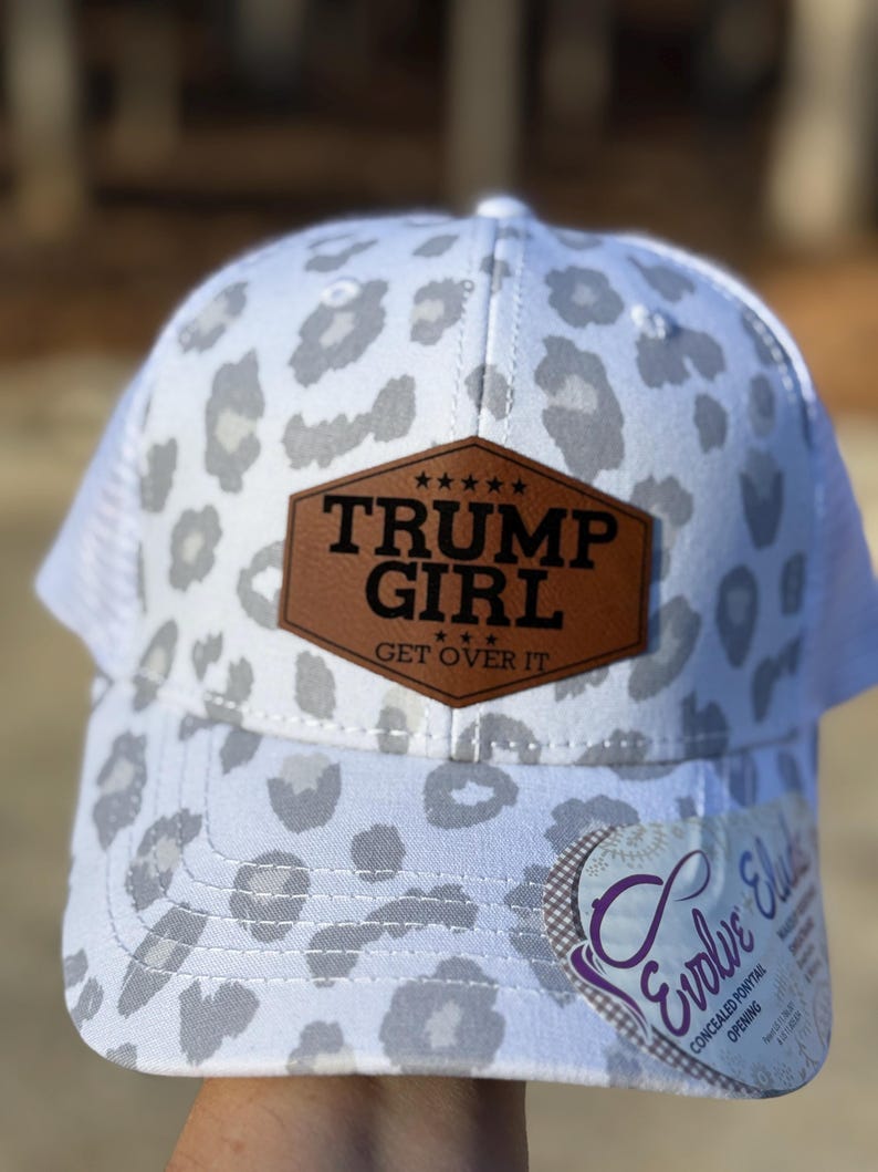 May include: White trucker hat with a grey leopard print pattern. The hat features a brown leather patch with the text "TRUMP GIRL GET OVER IT". The hat has a mesh back and a curved brim.