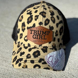 May include: A baseball cap with a leopard print design in tan and black, and a black mesh back. The cap has a brown leather patch with the words "TRUMP GIRL" and "GET OVER IT". A white tag is attached.
