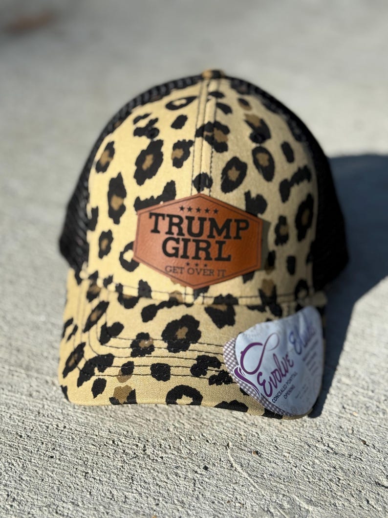 May include: A baseball cap with a leopard print design in tan and black, and a black mesh back. The cap has a brown leather patch with the words "TRUMP GIRL" and "GET OVER IT". A white tag is attached.