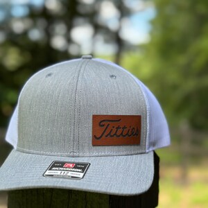 May include: A gray and white trucker hat with a brown leather patch that says "Titties" in a cursive font. The hat is made by Richardson 112.