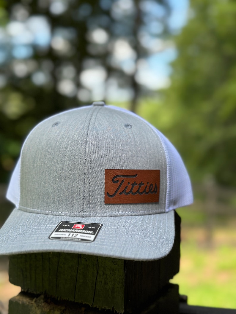 May include: A gray and white trucker hat with a brown leather patch that says "Titties" in a cursive font. The hat is made by Richardson 112.