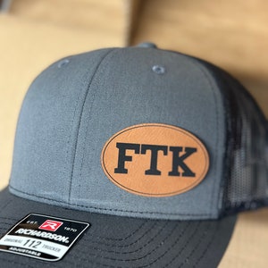 May include: A gray and black Richardson 112 trucker hat with a leather patch that reads "FTK". The hat has a mesh back and an adjustable snap closure. The patch is oval-shaped and has the letters "FTK" in a bold font.