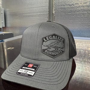 May include: Gray trucker hat with a black mesh back and a curved brim. The hat features the text "LEGALIZE BURNOUTS" in a black graphic on the front. A black and white tag reads "RICHARDSON ORIGINAL 112 TRUCKER ADJUSTABLE".