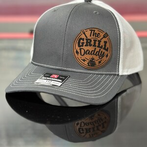 May include: A grey and white trucker hat with a brown leather patch that says "The Grill Daddy" in black lettering. The patch has a grill, spatula, and flames.