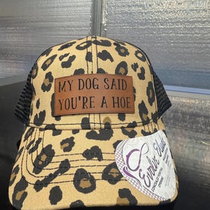 May include: A leopard print baseball cap with a black mesh back. A brown rectangular patch on the front features the black text MY DOG SAID YOU'RE A HOE. A white sticker on the brim displays the Evolve Elude logo and text regarding a ponytail opening.