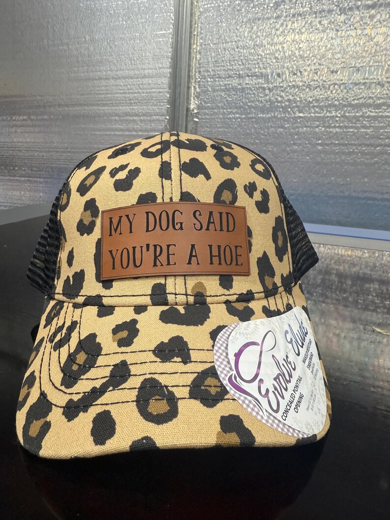 May include: A leopard print baseball cap with a black mesh back. A brown rectangular patch on the front features the black text MY DOG SAID YOU'RE A HOE. A white sticker on the brim displays the Evolve Elude logo and text regarding a ponytail opening.