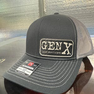 May include: A black and gray trucker hat with the text "GEN X I just don't care" on a rectangular patch. The hat has a curved brim with white stitching and an adjustable strap.