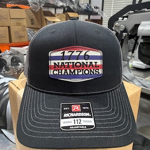 Peut inclure: Casquette de camionneur noire avec un dos en filet gris et une visière incurvée. L'avant présente un écusson avec "1776 NATIONAL CHAMPIONS" en rouge, blanc et bleu. Le chapeau comprend une étiquette Richardson et est réglable.