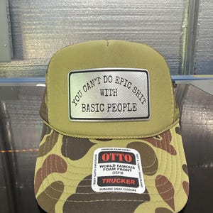 May include: A trucker hat with a camouflage pattern on the brim and a khaki-colored top. The hat features the text "YOU CAN'T DO EPIC SHIT WITH BASIC PEOPLE" on a silver patch. The hat also has the "OTTO" brand label.