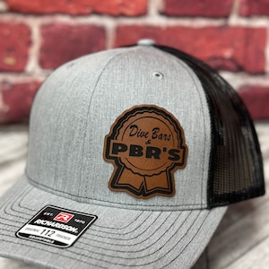 May include: Gray and black trucker hat with a brown leather patch that says "Dive Bars & PBR's". The patch is shaped like a ribbon. The hat has a black mesh back and an adjustable strap.