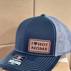 May include: A black and gray trucker hat with a brown leather patch that says "I ♥ HOT MOMS".