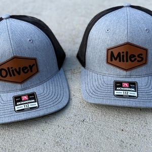 May include: Two gray and black trucker hats with brown leather patches. The patches are hexagonal and have the names "Oliver" and "Miles" written in black.