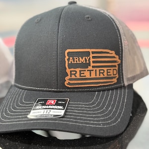 May include: A black and grey trucker hat with a brown leather patch that says "Army Retired" with a stylized American flag design. The hat is adjustable and has a Richardson logo on the back.