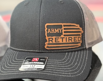May include: A black and grey trucker hat with a brown leather patch that says "Army Retired" with a stylized American flag design. The hat is adjustable and has a Richardson logo on the back.