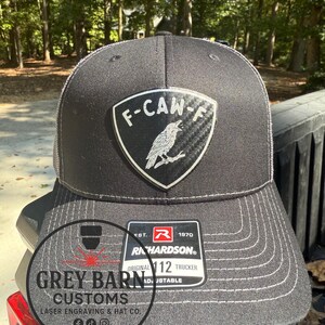 May include: Black trucker hat with a white stitched brim and a patch that reads "F-CAW-F" with a bird graphic. The hat has a Richardson tag that says "Original 112 Trucker". The hat is from Grey Barn Customs.