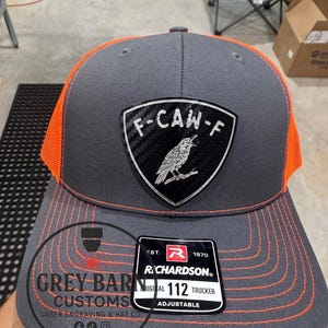 May include: A gray and orange trucker hat with a patch that reads "F-CAW-F" and a bird illustration. The hat has orange stitching and a mesh back. The tag reads "RICHARDSON 112 TRUCKER ADJUSTABLE".