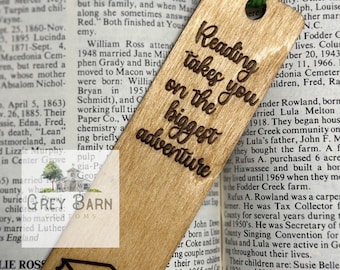 May include: Wooden bookmark with a brown background and the text "Reading takes you on the biggest adventure" engraved in a darker brown. The bookmark features a small illustration of two stacked books in the bottom left corner.