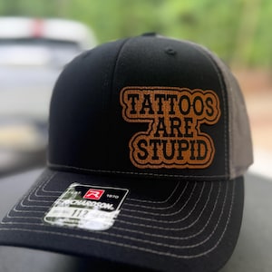 May include: Black and grey trucker hat featuring the text "TATTOOS ARE STUPID" in a brown, outlined font. The hat has a curved peak and a mesh back. A small tag with the brand name "RICHARDSON" is visible on the peak.