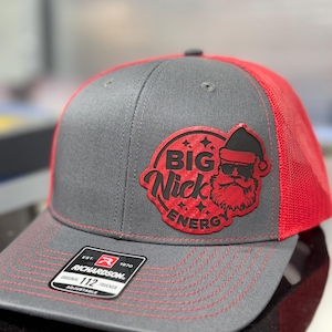 May include: Gray and red trucker hat with the text "BIG Nick ENERGY" and a Santa Claus graphic. The hat has a gray front panel, a red mesh back, and a curved brim. The Richardson 112 trucker hat is adjustable.
