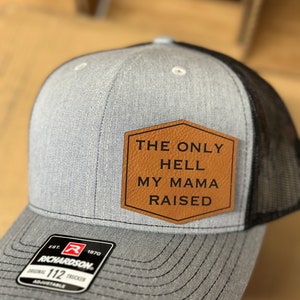 May include: A gray and black trucker hat with a brown leather patch that says "The Only Hell My Mama Raised". The hat has a Richardson logo on the side.