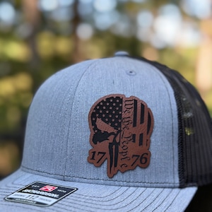 May include: A gray and black trucker hat with a brown leather patch featuring a skull with an American flag design and the text "We the People 1776".
