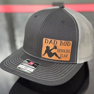 May include: A black and grey trucker hat with a brown leather patch that says "Dad Bod Drinking Team". The patch features a silhouette of a person holding a beer.