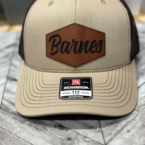 May include: A tan and black trucker hat with a brown leather patch that says "Barnes" in black lettering. The hat has a black mesh back and a Richardson 112 adjustable label.