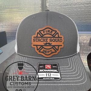 May include: A gray Richardson 112 trucker hat with a white mesh back. The hat features a brown leather patch with the words "FIRE STACHE SQUAD DEPT." The hat is adjustable and has white stitching.