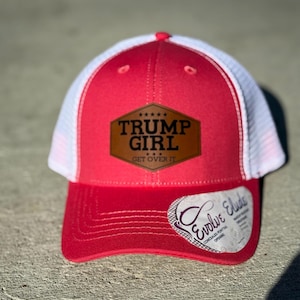May include: A red and white trucker hat featuring the text "TRUMP GIRL GET OVER IT" on a brown patch. The hat has a curved peak and a white mesh back. A white tag with the text "Evolve Elude" is attached to the peak.