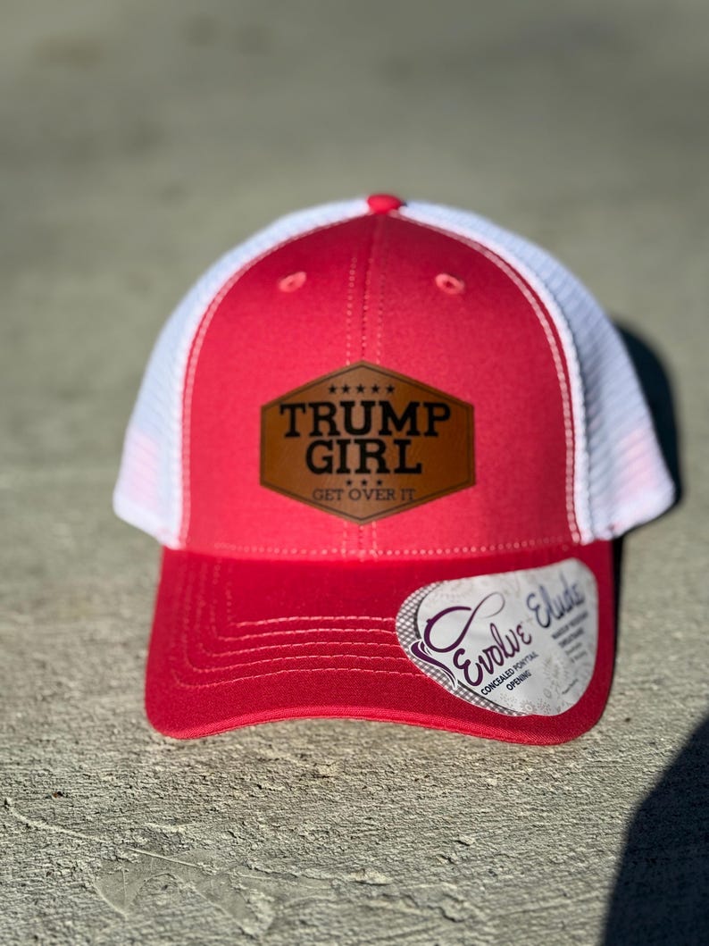 May include: A red and white trucker hat featuring the text "TRUMP GIRL GET OVER IT" on a brown patch. The hat has a curved peak and a white mesh back. A white tag with the text "Evolve Elude" is attached to the peak.