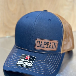 May include: A navy blue trucker hat with a brown mesh back. The hat has a brown leather patch on the front that says "CAPTAIN". The hat is adjustable and has a red and black label that says "EST. 1970 RICHARDSON. ORIGINAL 112 TRUCKER ADJUSTABLE".