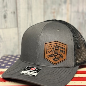 May include: A grey trucker cap with a black mesh back. It features a brown leather patch with a welder design and American flag elements. The cap has a curved peak and an adjustable strap.