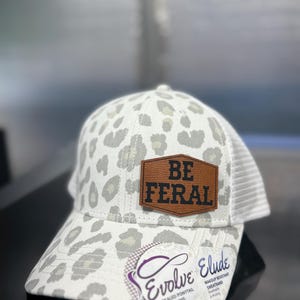 May include: White and gray leopard print baseball cap with a brown leather patch that reads "BE FERAL". The cap has a mesh back and a curved brim. The tag reads "Evolve Elude Concealed Ponytail Opening".
