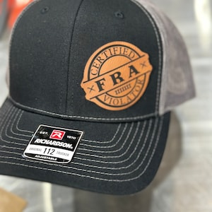 May include: Black and gray trucker hat with a brown leather patch that says "Certified FRA Violator". The hat is adjustable and has a Richardson 112 logo.