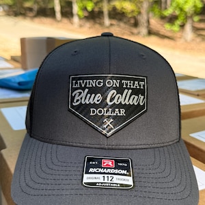 May include: A charcoal grey trucker cap with a black mesh back. The front displays a patch with the text "LIVING ON THAT Blue Collar DOLLAR" and a crossed hammer and wrench graphic. The cap has a curved peak and an adjustable strap.
