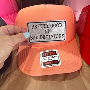 May include: An orange trucker hat with the text "PRETTY GOOD AT BAD DECISIONS" on a silver patch. The hat has a foam front and a snap closure. Other hats in pink, red, and black are visible in the background.