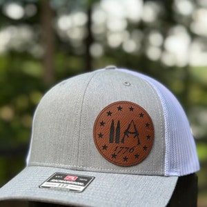 May include: A gray and white trucker hat with a brown leather patch featuring three bullets, a rifle, and the text "1776" surrounded by stars.