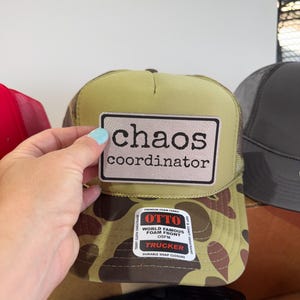 May include: A green and brown camouflage trucker hat with a white patch that reads "chaos coordinator." The hat has a mesh back and a snap closure. The brand is Otto.