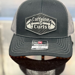 May include: Black and gray trucker hat with a mesh back and a curved brim. The hat features a patch with the words "Caffeine & Curls" and a barbell design. The Richardson brand label is visible on the brim.