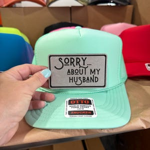 May include: A mint green trucker hat with a mesh back and a curved peak. The hat has a rectangular patch with the text "SORRY... ABOUT MY HUSBAND". Other hats in various colours are visible in the background.