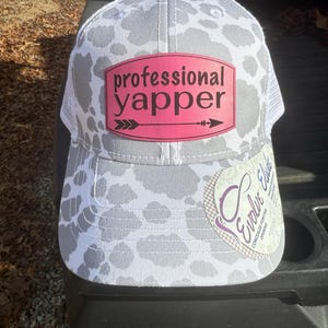 May include: A gray and white trucker hat with a cow print design. The hat features a pink patch with the words "professional yapper" and an arrow graphic. A white mesh back and a curved brim complete the design.