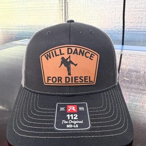 May include: A black trucker hat with a tan leather patch featuring a silhouette of a person dancing on a pole and the text WILL DANCE FOR DIESEL. A black sticker on the brim displays the text EST. 1970 R 112 The Original MD-LG.