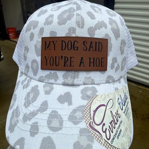 May include: White and gray leopard print baseball cap with a brown leather patch that reads "MY DOG SAID YOU'RE A HOE". The cap has a white mesh back and a curved brim. A tag with the text "Evolve Elude" is attached.