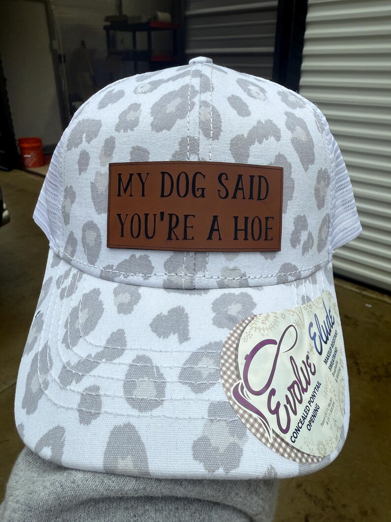 May include: White and gray leopard print baseball cap with a brown leather patch that reads "MY DOG SAID YOU'RE A HOE". The cap has a white mesh back and a curved brim. A tag with the text "Evolve Elude" is attached.