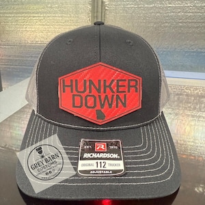 May include: Black trucker hat featuring a red and black hexagon patch with the text "HUNKER DOWN" and a Georgia outline. The hat has a grey mesh back and a curved peak. It is adjustable and has a Richardson tag.