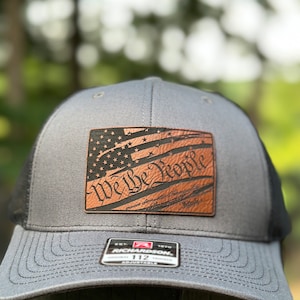 Peut inclure: Un chapeau de camionneur gris et noir avec un patch en cuir brun représentant un drapeau américain stylisé et le texte "We the People".