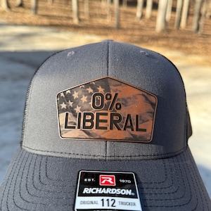 May include: Grey trucker hat with a black mesh back and a brown leather patch. The patch reads "0% LIBERAL" with a stars and stripes design. The hat has a Richardson tag.