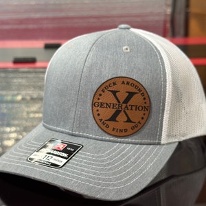 May include: A gray and white trucker hat with a leather patch. The patch features the text "FUCK AROUND AND FIND OUT" around the words "GENERATION X". The hat has a curved brim and an adjustable snap closure.