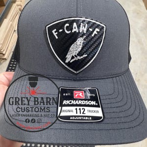May include: A gray Richardson 112 trucker hat with a black mesh back. The front features a shield-shaped patch with the text "F-CAW-F" and a bird illustration. The hat also has a "Grey Barn Customs" logo.