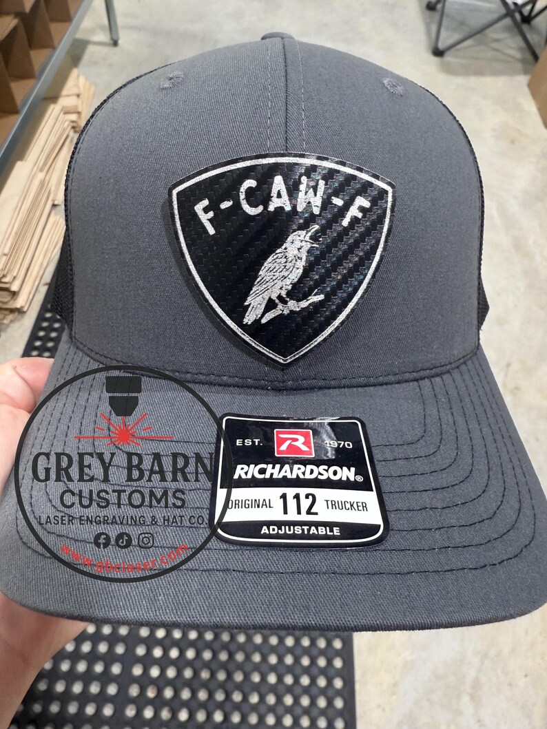 May include: A gray Richardson 112 trucker hat with a black mesh back. The front features a shield-shaped patch with the text "F-CAW-F" and a bird illustration. The hat also has a "Grey Barn Customs" logo.